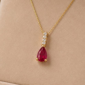 Può includere: Una collana in oro con un ciondolo a forma di pera con pietra rossa, sospeso a una catenina sottile. Il ciondolo presenta una piccola barra verticale incastonata con tre pietre chiare sopra la pietra centrale rossa a forma di goccia.