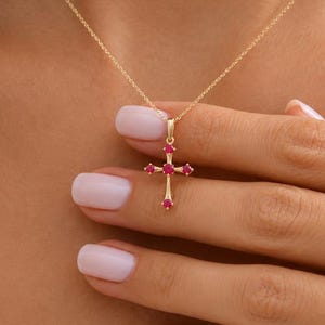 Puede incluir: Un collar de cadena de oro con un colgante de cruz engastado con cinco pequeñas piedras preciosas redondas de color rojo rosado vibrante. La cruz tiene un acabado en oro pulido y la delicada cadena descansa sobre la clavícula, resaltando el detalle.