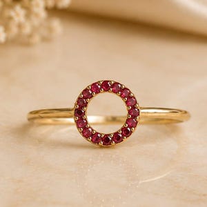 May include: A gold ring featuring a circular centerpiece adorned with small, vibrant red gemstones set in a continuous loop. The band is thin and polished, showcasing a simple design that highlights the circular arrangement of the red stones.