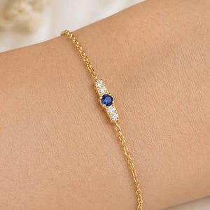 Könnte beinhalten: Ein zartes Goldkettenarmband mit einem horizontalen Mittelstück. Das Design besteht aus einem einzelnen runden blauen Edelstein, der von zwei kleinen klaren weißen Steinen auf jeder Seite flankiert wird und in einer polierten Goldfassung sitzt.