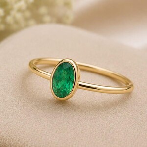 Peut inclure: Une bague en or ornée d'une pierre précieuse verte de forme ovale, sertie dans une monture lisse. L'anneau est fin et poli, présentant un design épuré qui met en valeur la couleur vive de la pierre centrale sur la finition métallique.
