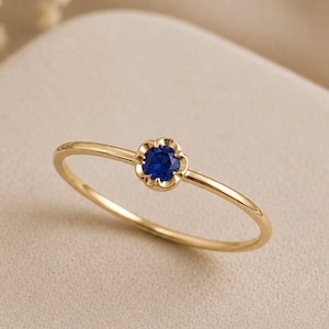 May include: A thin gold ring featuring a small, round, deep blue gemstone set within a delicate, flower-shaped metal frame. The band is smooth and polished, showcasing a minimalist design that highlights the vibrant contrast between the gold and blue.