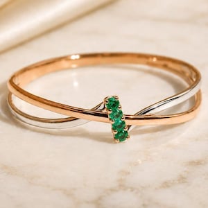 Puede incluir: Un brazalete bicolor en oro y plata. La banda dorada cruza la banda plateada, con una fila vertical de piedras preciosas verdes en el centro. El brazalete se exhibe sobre una superficie de mármol.