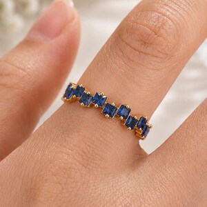 May include: A gold band ring featuring a series of small, square-cut deep blue gemstones set in a continuous, slightly staggered row along the top of the band, creating a textured and geometric appearance across the finger.