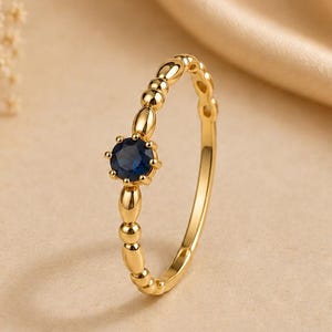 May include: A gold ring with a dark blue gemstone center. The ring band features a beaded design. The ring is set against a soft, neutral background with a hint of floral accents.