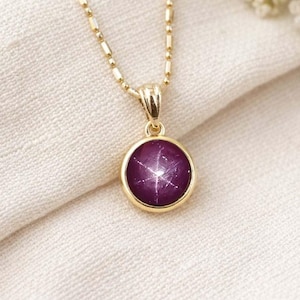 Op de afbeelding: Een gouden ketting met een ronde, dieppaarse hanger. De hanger heeft een stervormig patroon en is gezet in een gouden bezel. De ketting is een delicate gouden kralenketting en de ketting rust op een witte stof.