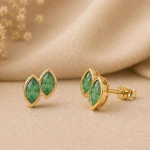 Puede incluir: Pendientes de oro con piedras preciosas verdes esmeralda con forma de marquesa. Los pendientes tienen un diseño en forma de hoja, con dos piedras preciosas por pendiente. Se muestran sobre una tela beige.