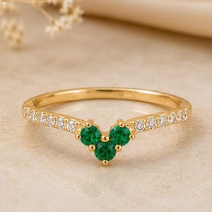 Op de afbeelding: Een goudkleurige ring met een V-vormig ontwerp. De ring heeft drie ronde, heldergroene edelstenen in het midden en kleine, heldere edelstenen langs de band. De ring is geplaatst tegen een neutrale achtergrond.