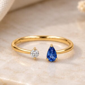 Puede incluir: Un anillo abierto de oro con una piedra redonda de talla brillante en un extremo y un zafiro azul en forma de pera en el otro. La banda es fina y pulida, mostrando un diseño sencillo con dos piedras preciosas engastadas en garras de oro.