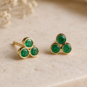 May include: A pair of stud earrings featuring three small, round, vibrant green stones set in a triangular cluster of polished yellow gold. Each stone is securely held in a bezel setting, creating a cohesive, floral-inspired shape for each earring.