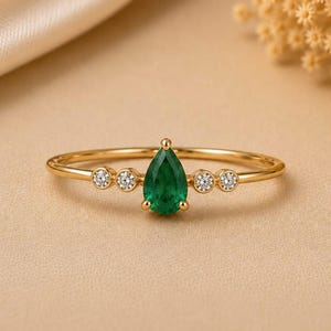 May include: A gold ring featuring a central pear-shaped green gemstone flanked by two small round clear stones on each side. The band is thin and polished, showcasing a delicate design that highlights the vibrant green hue of the pear-cut stone.