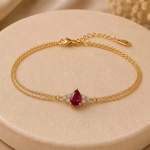 Könnte beinhalten: Ein goldfarbenes Doppelkettenarmband mit einem zentralen birnenförmigen roten Edelstein, der auf jeder Seite von zwei kleinen klaren Steinen flankiert wird. Die feine Metallkette verfügt über einen Karabinerverschluss und ein Verlängerungskettchen.