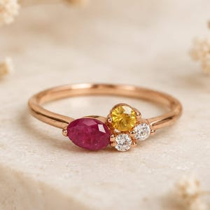 Peut inclure: Une bague en or rose ornée d'un groupe de pierres précieuses. La bague présente un rubis en forme de marquise, un groupe de pierres jaunes et deux petites pierres blanches. La bague est posée sur une surface blanche.