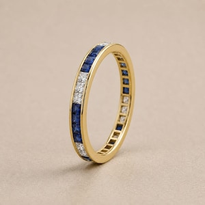 Peut inclure: Une bague en or avec des saphirs bleus et des diamants blancs taillés en carré en alternance. La bague présente un motif continu de pierres précieuses serties dans un anneau en or jaune, de style éternité.