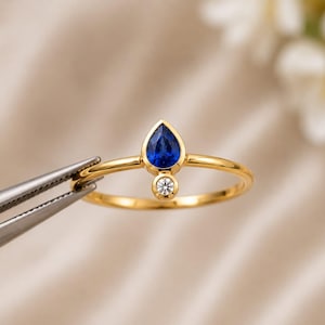 Op de afbeelding: Een gouden ring met een peervormige blauwe saffier in een zetting, geplaatst direct boven een kleine, ronde, helderwitte diamant die ook in een zetting is geplaatst. De ringband is dun, gepolijst en heeft een cirkelvormig ontwerp.