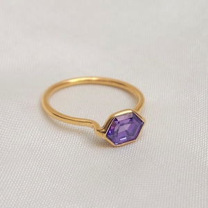 Peut inclure: Une bague en or ornée d'une pierre précieuse hexagonale violette. La bague a un anneau fin et un design minimaliste. La pierre précieuse est d'une teinte violette vive.