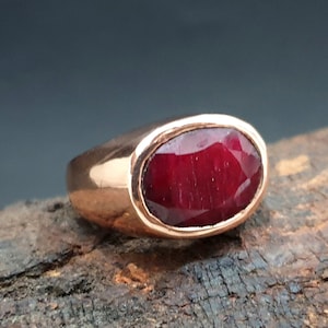 Pode incluir: Um anel de ouro rosa com uma grande gema de rubi de forma oval. O rubi é de cor vermelho escuro e tem um corte facetado.