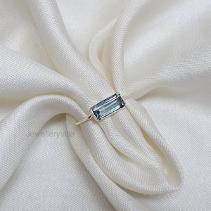 Op de afbeelding: Een zilveren ring met een lichtblauwe rechthoekige edelsteen. De ring ligt op een witte stoffen achtergrond.