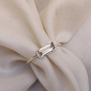 Pode incluir: Um anel de prata com uma gema retangular transparente. O anel está sobre um fundo de tecido branco.