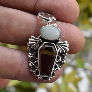 May include: Silver pendant featuring a coffin-shaped gemstone with a white opal at the top. The pendant is detailed with intricate silverwork.