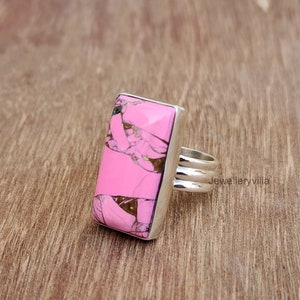 May include: A pink and brown rectangular gemstone ring with a silver band. The ring is set on a wooden surface.