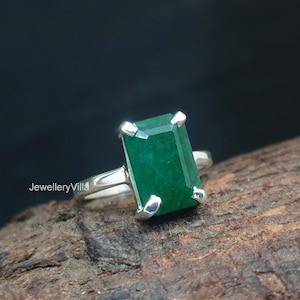 Puede incluir: Un anillo de plata con una gran piedra preciosa verde esmeralda. El anillo está colocado sobre una superficie de madera.
