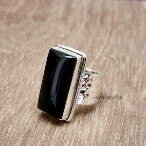 May include: A silver ring featuring a black rectangular gemstone. The ring band has a textured design and the words "Jewelleryvilla" are engraved on the side.