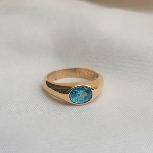 May include: A gold ring with a bright blue oval gemstone set in the center. The ring has a smooth, polished band and a classic design. The gemstone is a vibrant shade of blue, adding a pop of color to the ring.