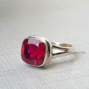 Pode incluir: Um anel de prata com uma grande pedra preciosa vermelha quadrada. O anel tem uma banda dupla e o texto "JewelleryVilla" é visível na banda.