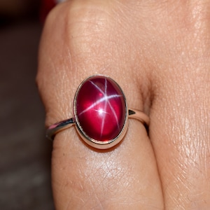Peut inclure: Une bague en argent avec une pierre précieuse en rubis étoilé rouge. Le rubis est de forme ovale et présente un motif en forme d'étoile au centre.
