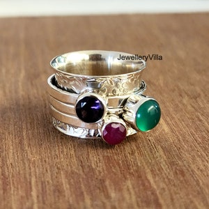 May include: A silver spinning ring featuring three gemstones: a purple amethyst, a green agate, and a red ruby. The ring is engraved with a floral design.