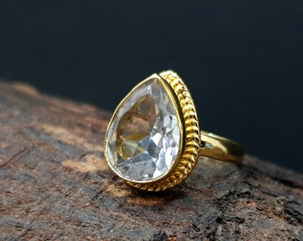 May include: A gold ring with a clear teardrop-shaped gemstone. The ring has a textured band.