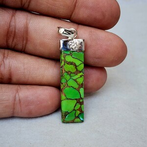 Pu&ograve; includere: Un ciondolo rettangolare con una vibrante pietra verde e marrone, sormontato da un'ansa color argento. La pietra presenta un motivo naturale unico. Il ciondolo &egrave; tenuto in una mano.