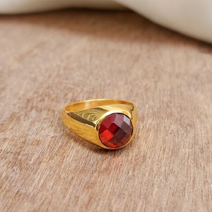 Könnte beinhalten: Ein goldener Ring mit einem großen, roten Edelstein. Der Ring liegt auf einer Holzoberfläche.