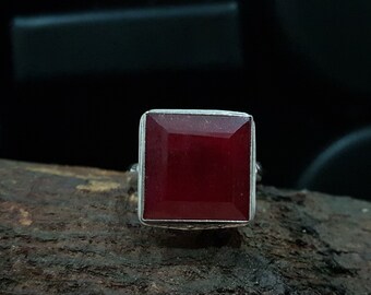 May include: A square-cut, deep red gemstone ring with a silver band. The gemstone is set in a square shape, and the ring is displayed on a textured wooden surface. The ring's design is simple and elegant, suitable for everyday wear.