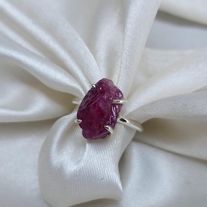 May include: A silver ring featuring a raw, uncut ruby gemstone. The ruby is a deep, vibrant red color and is held in place by four silver prongs. The ring is set against a backdrop of white fabric.