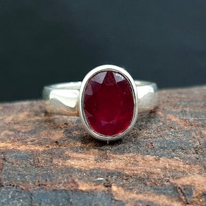 Puede incluir: Un anillo de plata con una gran piedra preciosa de rubí de forma ovalada. El rubí es de color rojo intenso y está engastado en una banda de plata sencilla.