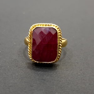 May include: A gold ring with a large, rectangular, faceted ruby gemstone set in a gold rope-like border.