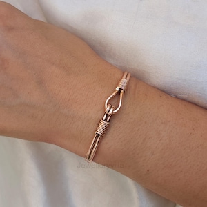 May include: A copper-colored bracelet with a hook-and-eye clasp. The bracelet features a simple, yet elegant design with wrapped wire detailing on the clasp and the ends of the bracelet. The jewelry is worn on a wrist.