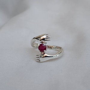 Peut inclure: Une bague en argent représentant deux mains étreignant une pierre précieuse rouge ronde. L'anneau de la bague épouse le doigt, les mains étant positionnées pour bercer la pierre précieuse. La bague est posée sur un fond blanc.