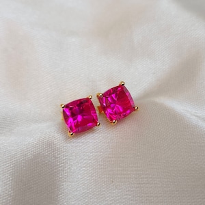 May include: A pair of square-cut, hot pink gemstone stud earrings with gold-coloured settings. The earrings are displayed on a white, textured surface, highlighting their vibrant colour and design.