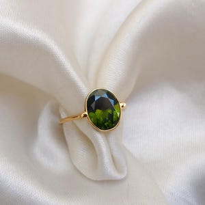 May include: A gold ring with an oval, green gemstone. The ring has a slender band and a simple design. The gemstone is set in a gold bezel. The ring is displayed on white, silky fabric.