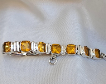 May include: A silver bracelet with a row of square-shaped amber gemstones. The bracelet has a clasp and a small dangling charm.