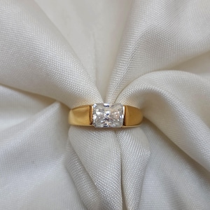 Puede incluir: Un anillo de oro con una gema rectangular y transparente. El anillo está colocado sobre una tela blanca texturizada. La banda del anillo es de color oro.