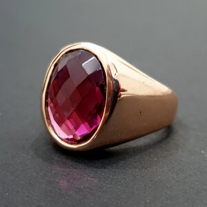 May include: A rose gold ring with a large, oval-shaped, faceted, dark pink gemstone.