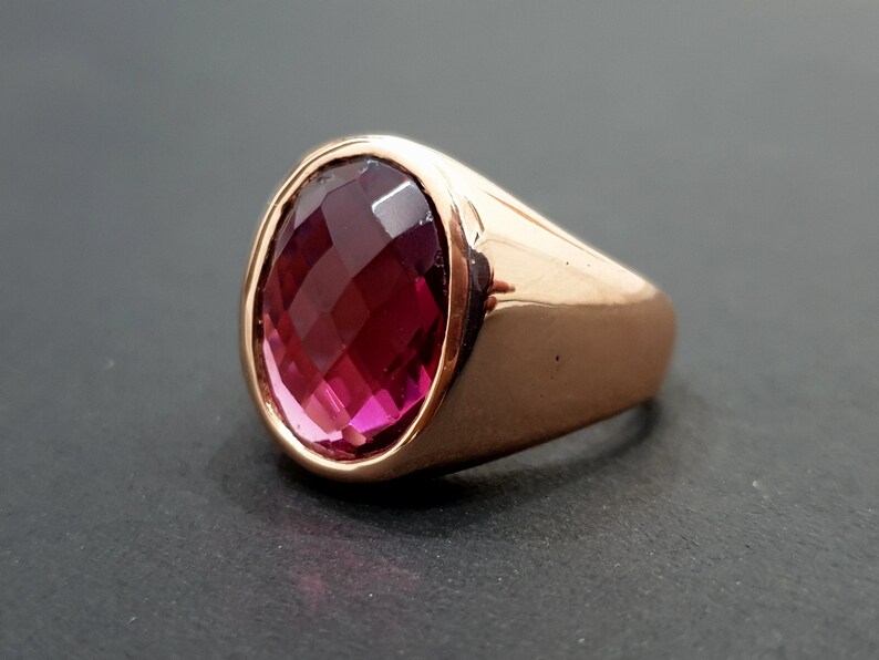 May include: A rose gold ring with a large, oval-shaped, faceted, dark pink gemstone.