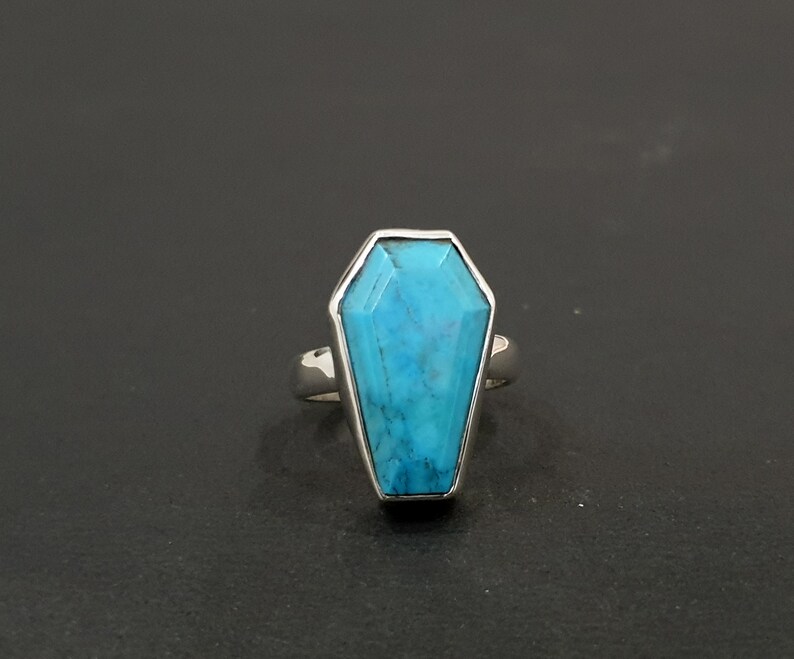 K&ouml;nnte beinhalten: Ein Silberring mit einem Sarg-f&ouml;rmigen T&uuml;rkisstein. Der hellblaue Stein ist facettiert und in einem polierten Silberband gefasst. Der Ring wird vor einem dunklen Hintergrund gezeigt.