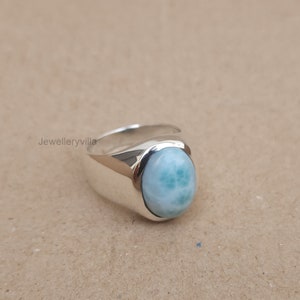 K&ouml;nnte beinhalten: Ein silberner Ring mit einem ovalen, blau-gr&uuml;nen Edelstein. Der Ring hat ein breites Band.