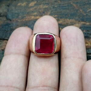 Pu&ograve; includere: Un anello in oro con una grande pietra preziosa rosso scuro a taglio quadrato, incastonata in una fascia di metallo liscia e lucida. La pietra ha una superficie sfaccettata che riflette la luce e la montatura &egrave; una semplice cornice in oro.