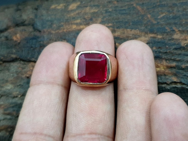 Pu&ograve; includere: Un anello in oro con una grande pietra preziosa rosso scuro a taglio quadrato, incastonata in una fascia di metallo liscia e lucida. La pietra ha una superficie sfaccettata che riflette la luce e la montatura &egrave; una semplice cornice in oro.
